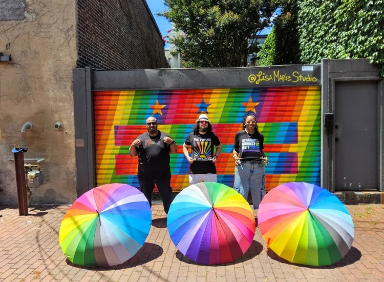Vista Supports team members standing in front of a colorful pride mural holding rainbow umbrellas, representing inclusion, diversity, and community support